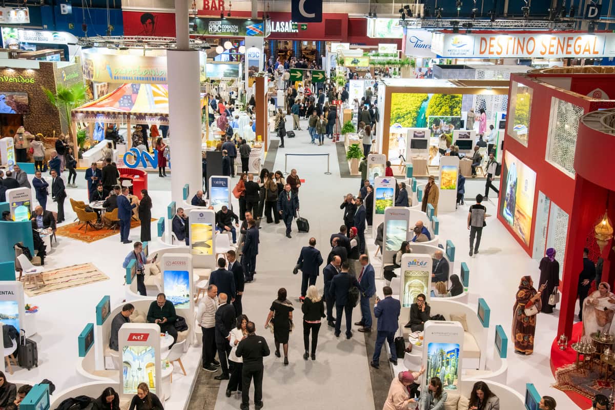 Een drukke overdekte expohal vol mensen die wandelen en praten tussen de stands en displays, met spandoeken en bewegwijzering voor verschillende landen en bestemmingen zichtbaar boven de exposities. Toerismebeurs FITUR in Madrid, 2025