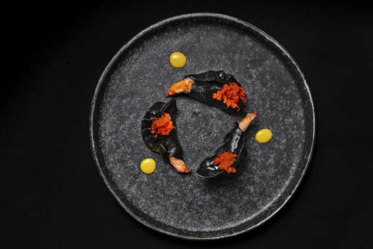 Zwarte dumplings met garnalen, tobiko-kaviaar en saus, geserveerd op een zwart bord tegen een zwarte achtergrond.