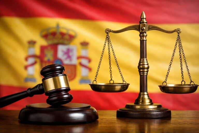 Rechtershamer en weegschaal van Justitia op tafel, met de Spaanse vlag onscherp op de achtergrond.