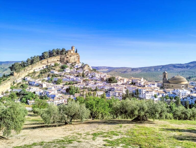 Een dorp op een heuvel met witte gebouwen, een kerk bovenop de heuvel, een gebouw met een koepel op de voorgrond en olijfbomen op de voorgrond onder een helderblauwe hemel. Montefrío Granada, een van de mooiste dorpen van Spanje.