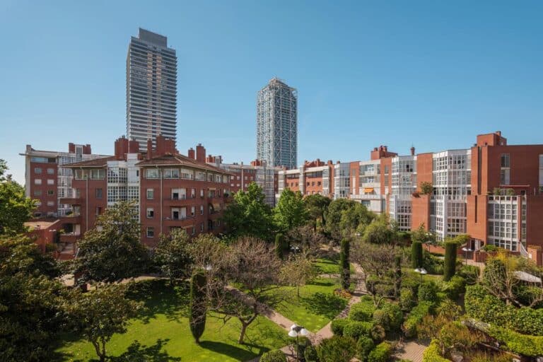 Modern wooncomplex in Barcelona met rode bakstenen appartementen, een groen park en twee glazen wolkenkrabbers op de achtergrond.