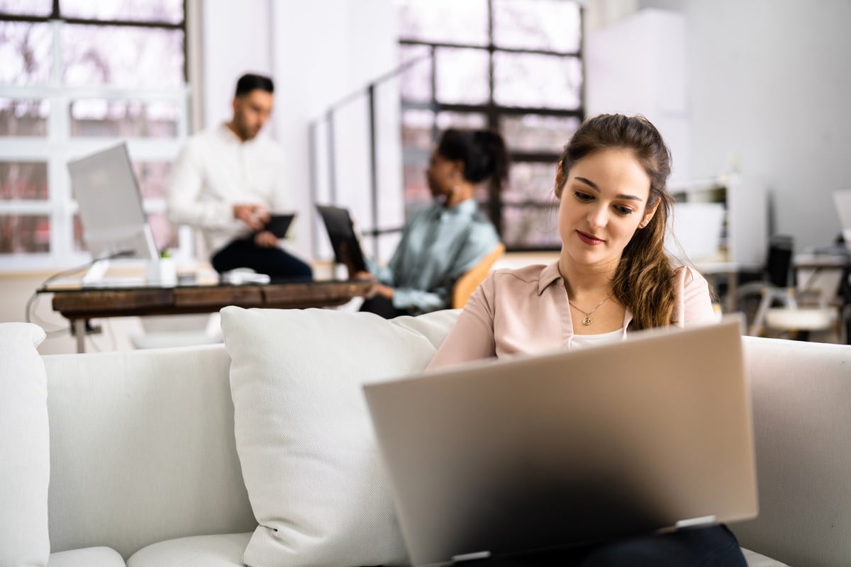 Jonge vrouw op een bank in een co-living ruimte met laptop op schoot, terwijl andere jonge bewoners op de achtergrond aanwezig zijn