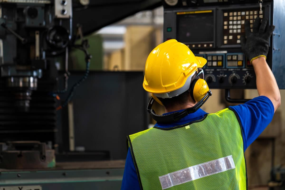 Arbeider met veiligheidshelm en gehoorbescherming bedient een industriële machine in een fabriekshal.