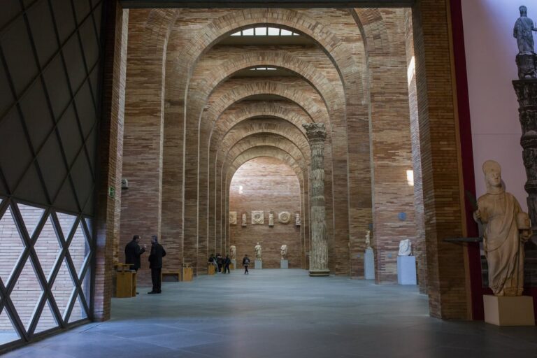 Interieur van een museumzaal met bakstenen bogen, Romeinse beelden en enkele bezoekers.