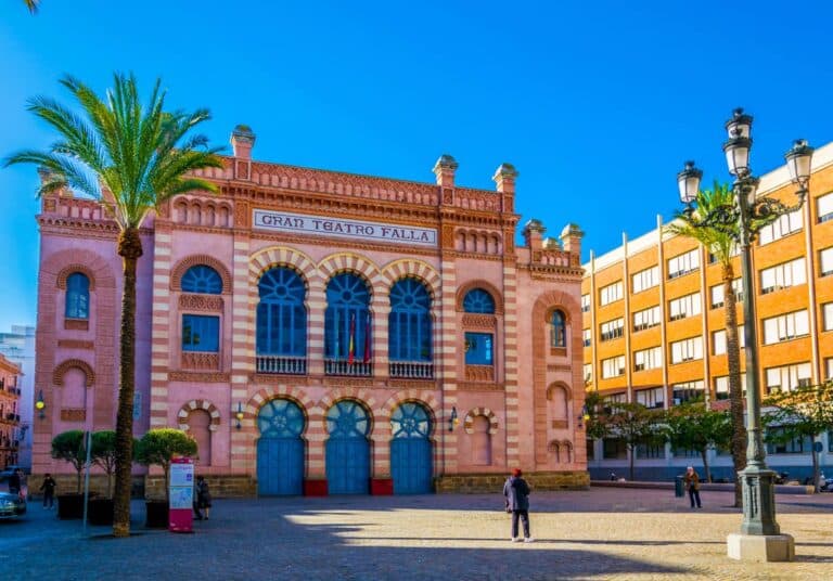 Voorgevel van het Gran Teatro Falla in Cádiz met rood-witte baksteen, bogen, palmbomen en een plein met enkele mensen