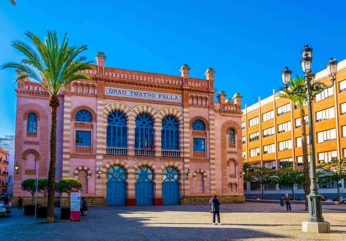 Voorgevel van het Gran Teatro Falla in Cádiz met rood-witte baksteen, bogen, palmbomen en een plein met enkele mensen