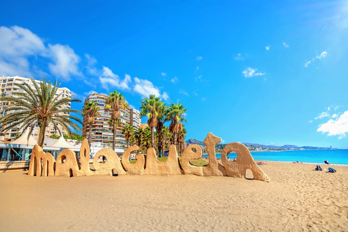 Het strand van Malagueta in Málaga met palmbomen, appartementen en het bekende Malagueta-bord aan zee onder een heldere blauwe lucht.