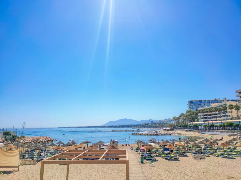 Zonnig strand aan de Costa del Sol met ligbedden, parasols, kalme zee en boulevard onder een heldere blauwe lucht.