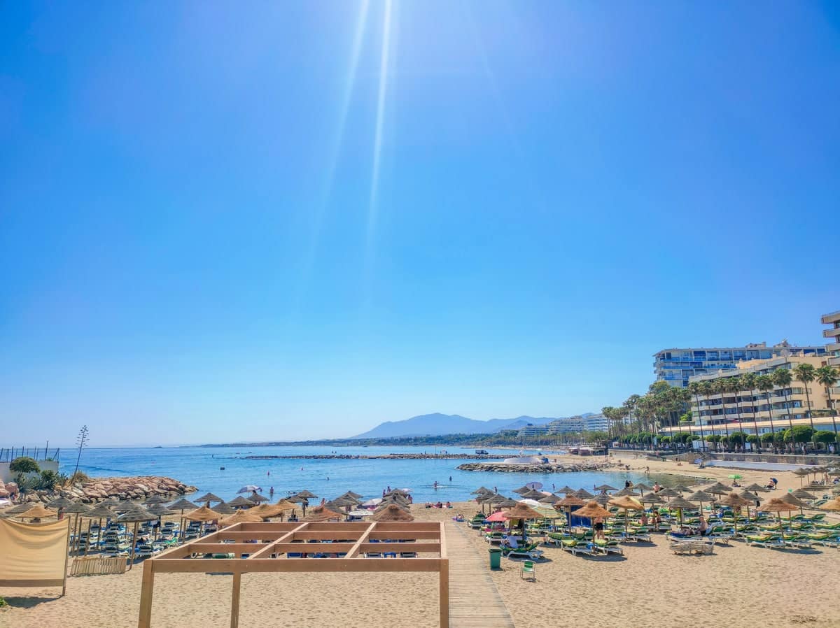 Zonnig strand aan de Costa del Sol met ligbedden, parasols, kalme zee en boulevard onder een heldere blauwe lucht.